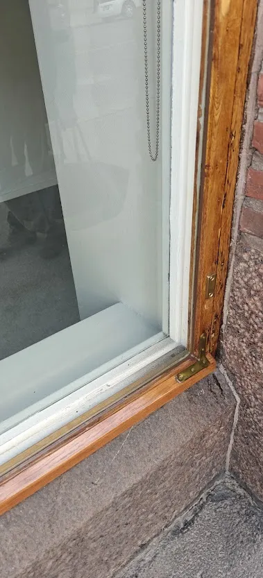 Lähikuva ikkunan alaosasta, jossa näkyy puinen ikkunanpoka, lasi, kivinen ikkunalauta ja metalliset kulmaraudat, jotka ovat tyypillisiä ikkunoiden kunnostuskohteita.