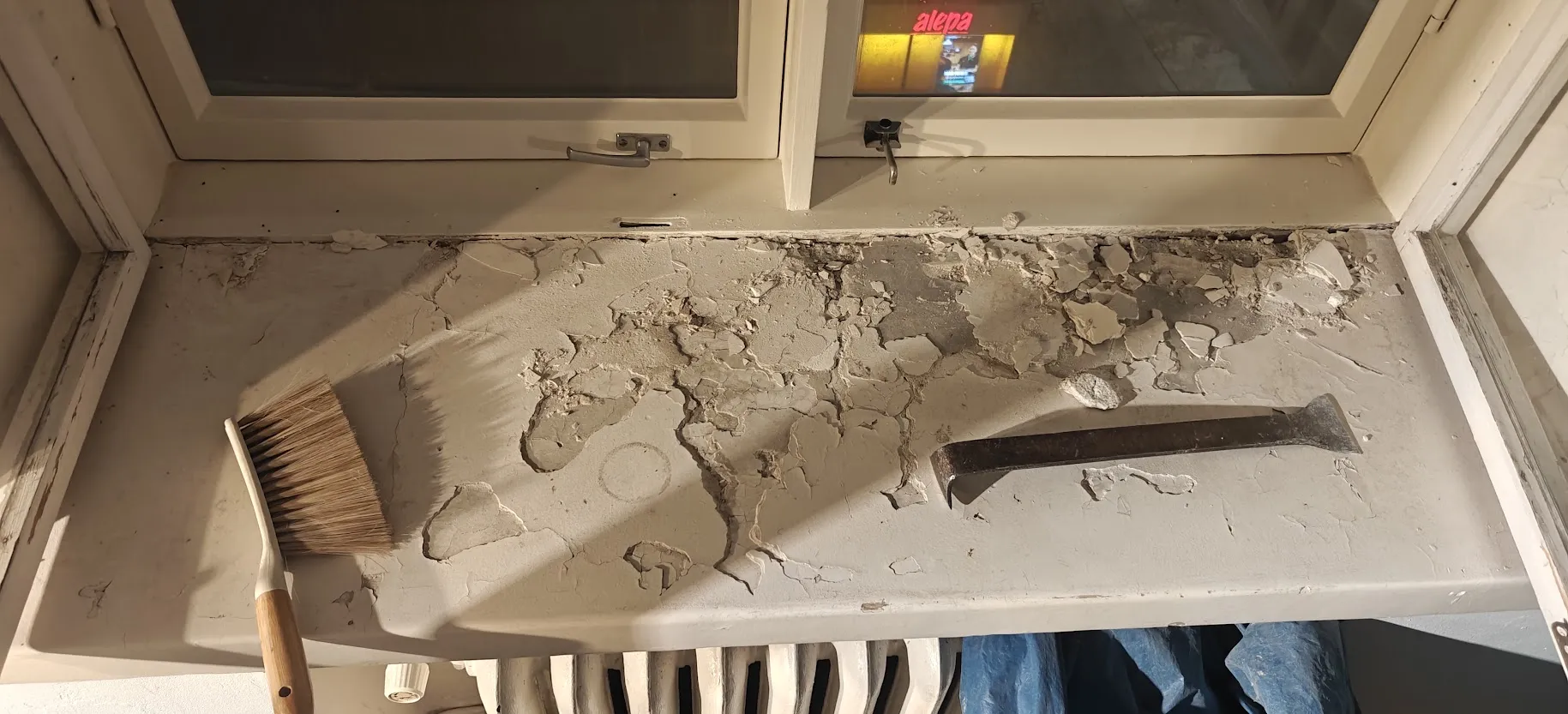 Ikkunan alla oleva seinäpinta, jossa maali hilseilee ja paljastaa kunnostusta vaativan pinnan, harja ja sorkkarauta ovat esillä.