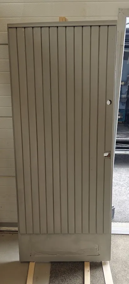 Harmaa pystypaneeliovi, jossa on lukon ja kahvan aukot, on tuettu puupalikoilla oven kunnostusta varten.