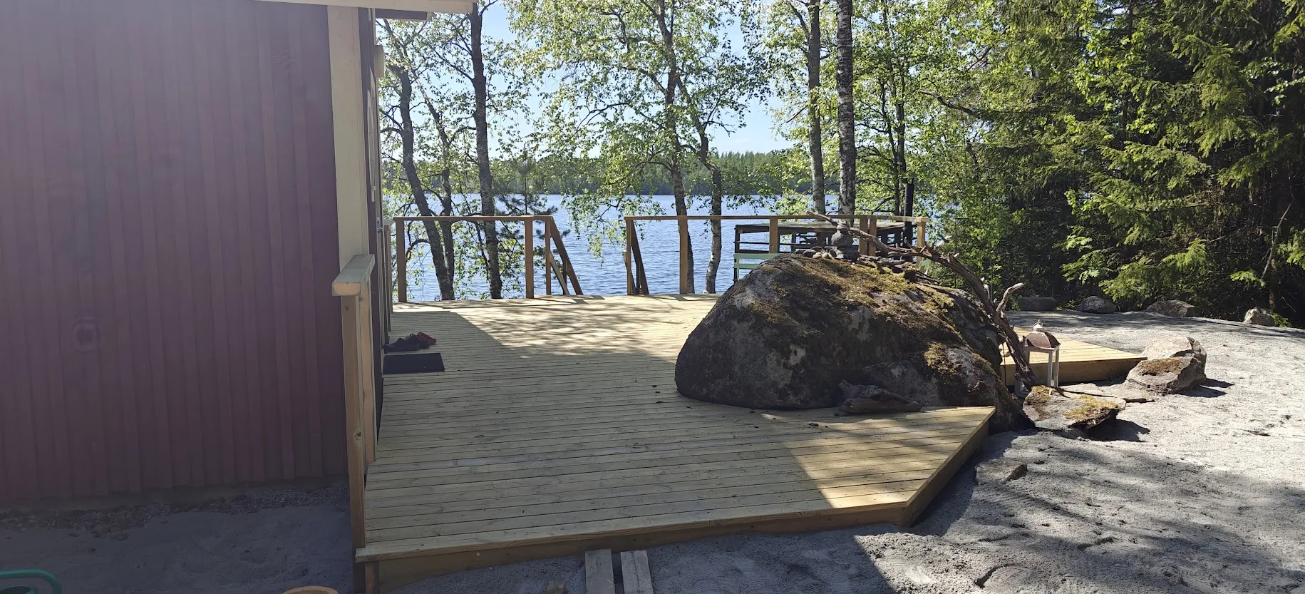 Uusi puinen terassi punaisen rakennuksen vieressä järven rannalla, suuren sammaleisen kiven ja puiden ympäröimänä.