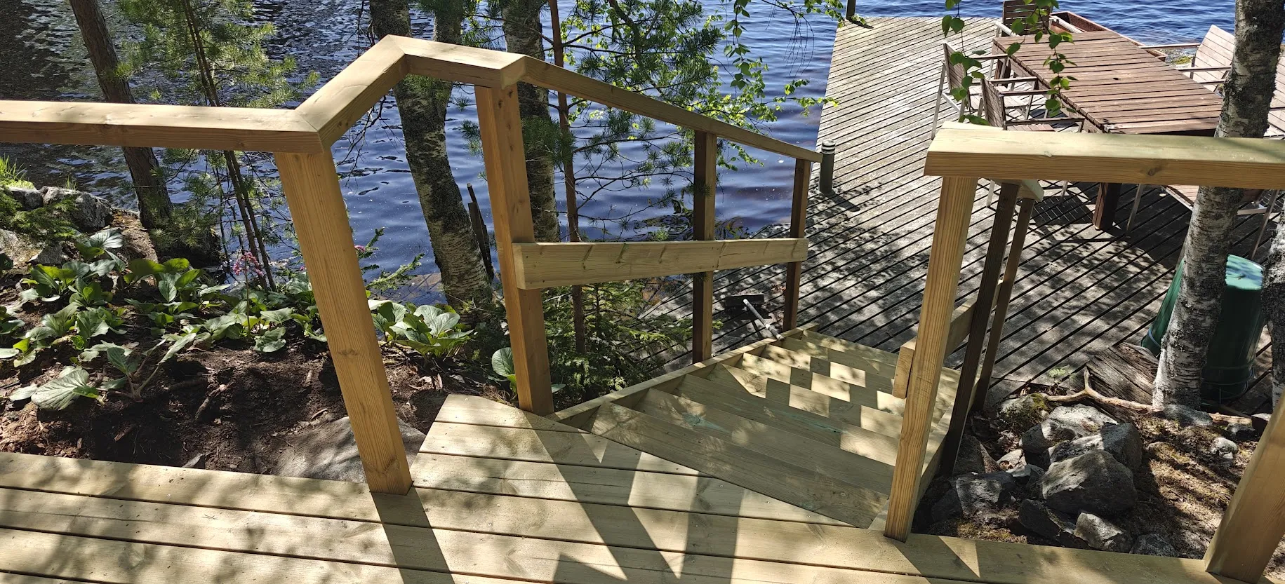 Puinen portaikko ja kaide laskeutuvat järven rantaan, jossa näkyy puinen laituri ja kunnostettu ranta-alue.