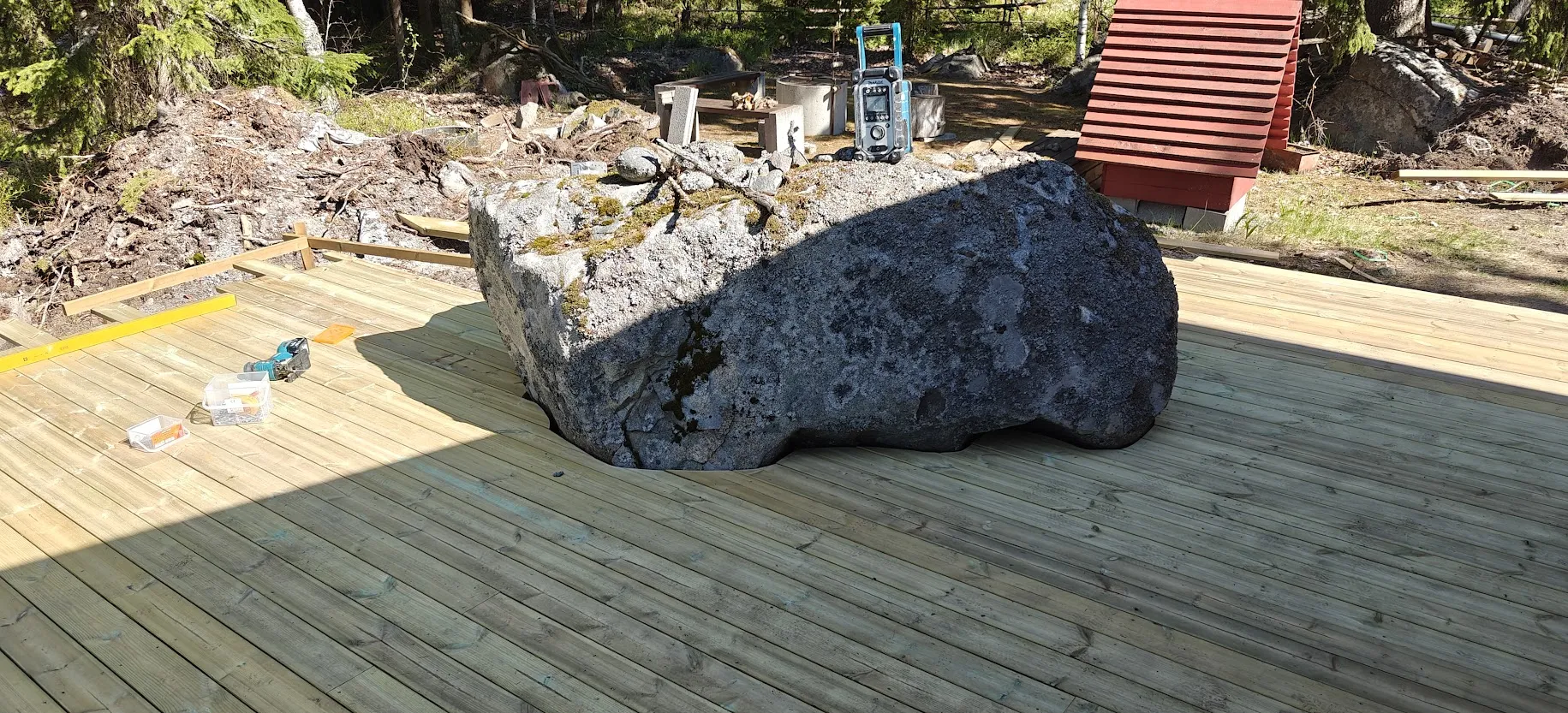 Uusi puinen terassi rakennetaan suuren kiven ympärille, ja remonttityökalut ovat esillä aurinkoisella säällä.
